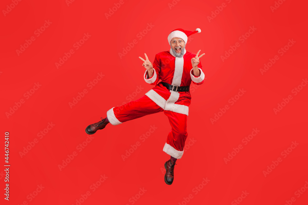 Full length portrait of happy elderly man with gray beard wearing santa claus costume jumping showing v gesture with fingers, celebrating winter holiday. Indoor studio shot isolated on red background. Full length portrait of happy elderly man with gray beard wearing santa claus costume jumping showing v gesture with fingers, celebrating winter holiday. Indoor studio shot isolated on red background.
