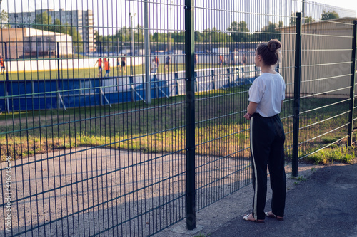 A girl stands and looks through the fence at the athletes playing at the stadium. The girl wants, but cannot play sports.