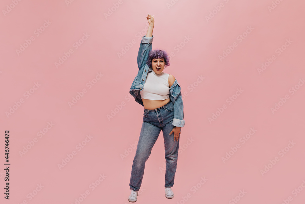 Obraz premium Charming woman in good mood with short hair in tight jeans and wide blue jacket shows her tongue at camera on pink background..