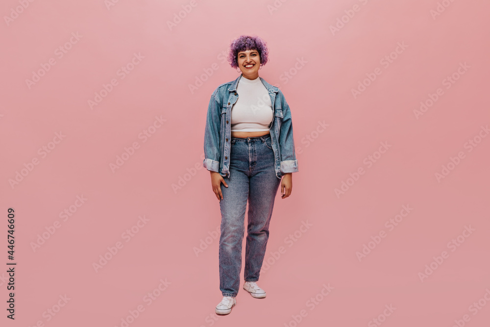 Full-length portrait of joyful woman with short purple hair in denim suit, white sneakers and light top smiling at camera..