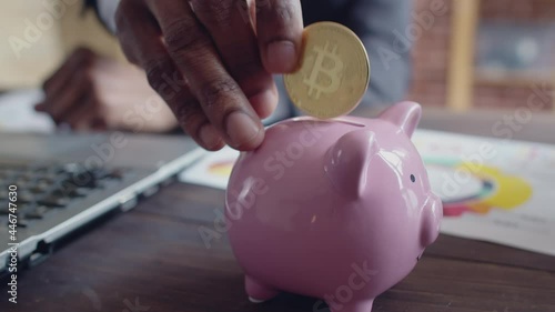 Black man businessman in suit and tie uses laptop and puts in the piggy bank on an office table a gold bitcoin coin, close-up. photo