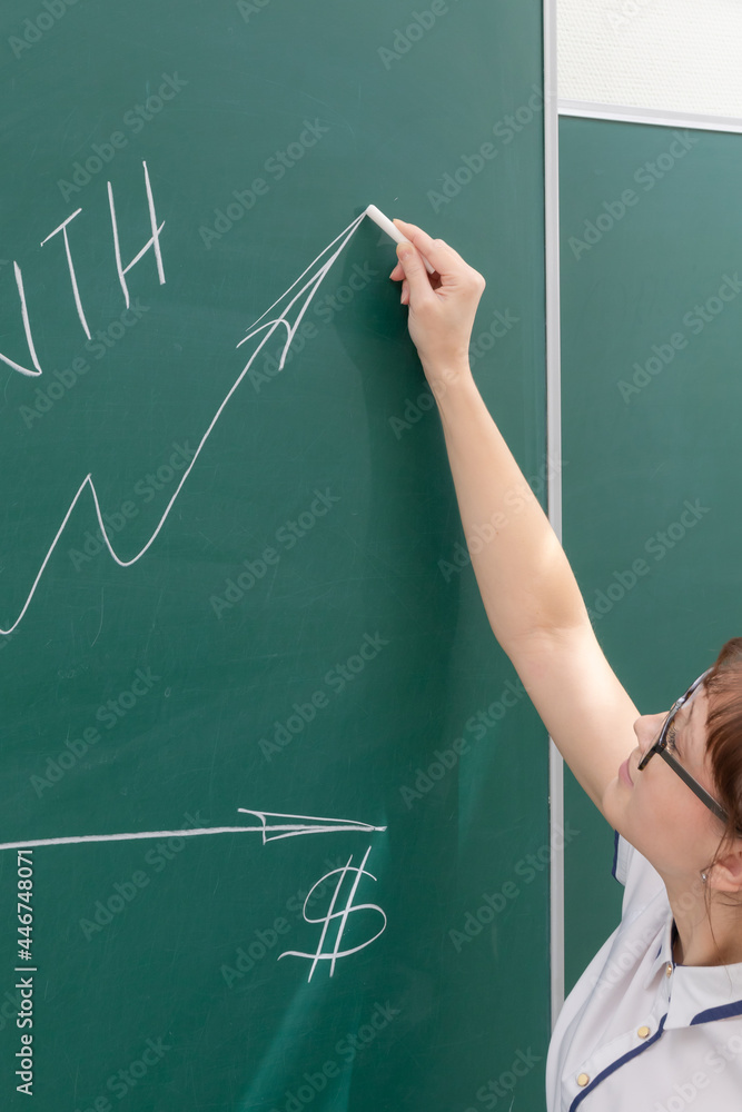 young woman business coach in a white blouse and glasses at the chalkboard in the office draws a profit graph. close-up