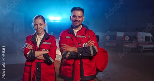 Portrait of happy Caucasian young man and woman paramedics at ambulance. Male and female doctors in red uniforms smiling at camera. Physicians and colleagues at night during coronavirus pandemic. photo