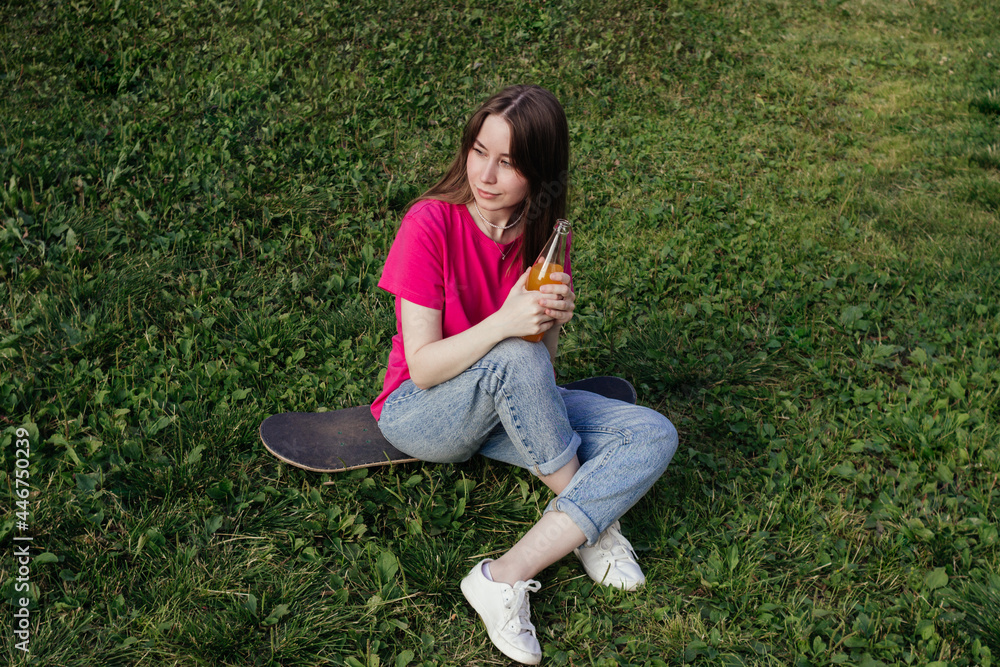 Smiling Young woman in pink t-shirt sitting on skateboard on green grass and holding bottle of lemonade. Summer activity, healthy lifestyle, sport, fitness. Female skater. Having fun outdoors Smiling Young woman in pink t-shirt sitting on skateboard on green grass and holding bottle of lemonade. Summer activity, healthy lifestyle, sport, fitness. Female skater. Having fun outdoors