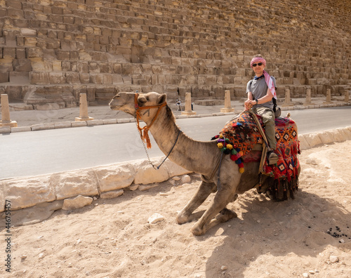 A tourist on a camel poses against the backdrop of the pyramids in Giza  Cairo  Egypt. Kufiya is wrapped around the man s head. Camel knelt down.
