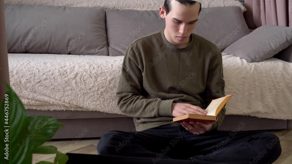 Muslim student reads a book at home sitting on the floor. Traditional Religious Education School, Distance learning, lockdown. Young man teenager is studying Muslim student reads a book at home sitting on the floor. Traditional Religious Education School, Distance learning, lockdown. Young man teenager is studying