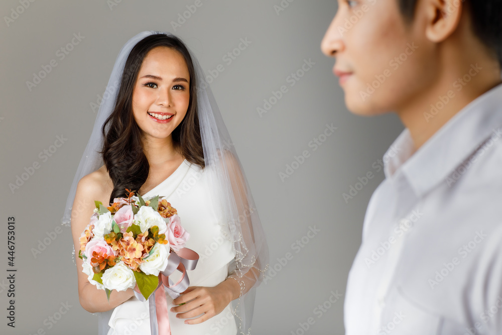 Young attractive Asian couple, selective focus on bride wearing wedding gown with veil holding bouquet of flower. Blurry man wearing white shirt on foreground. Concept for pre wedding photography Young attractive Asian couple, selective focus on bride wearing wedding gown with veil holding bouquet of flower. Blurry man wearing white shirt on foreground. Concept for pre wedding photography