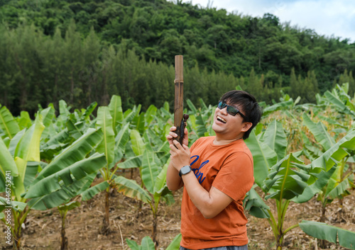 Asian man laughing with happiness holding bamboo bong in banana trees farm on green hill, bong use for marijuana or nicotinic smoking, narcotics addicted concept photo