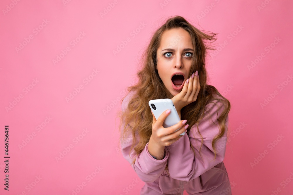 Photo of attractive crazy amazed surprised young woman wearing casual stylish clothes standing isolated over background with copy space holding and using mobile phone looking at camera Photo of attractive crazy amazed surprised young woman wearing casual stylish clothes standing isolated over background with copy space holding and using mobile phone looking at camera