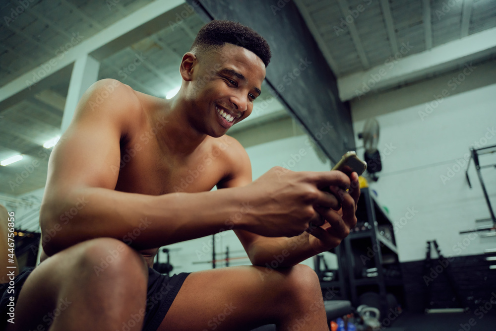 Young African American male looking at his phone to reply to an online message. Happy, mixed race male smiling at his phone while sitting down in the gym. High quality photo Young African American male looking at his phone to reply to an online message. Happy, mixed race male smiling at his phone while sitting down in the gym. High quality photo