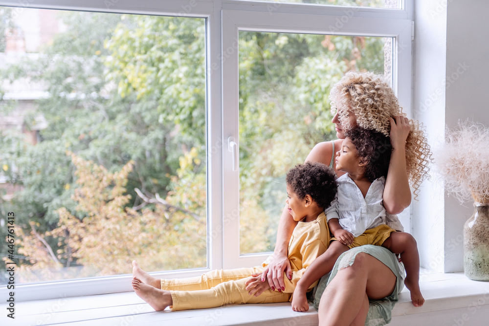 Young blond Caucasian woman hugging African American children, daughter and son. Happy family is sitting on wide windowsill, looking out the window. Single Parenting. Adoption mixed race. Multi ethnic