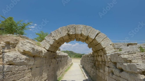 Olympia, Greece POV at Archaeological site area that hosted ancient sports Games. Walking to the stone built arch and entrance of the stadium of athletic contests on classical times, against blue sky.