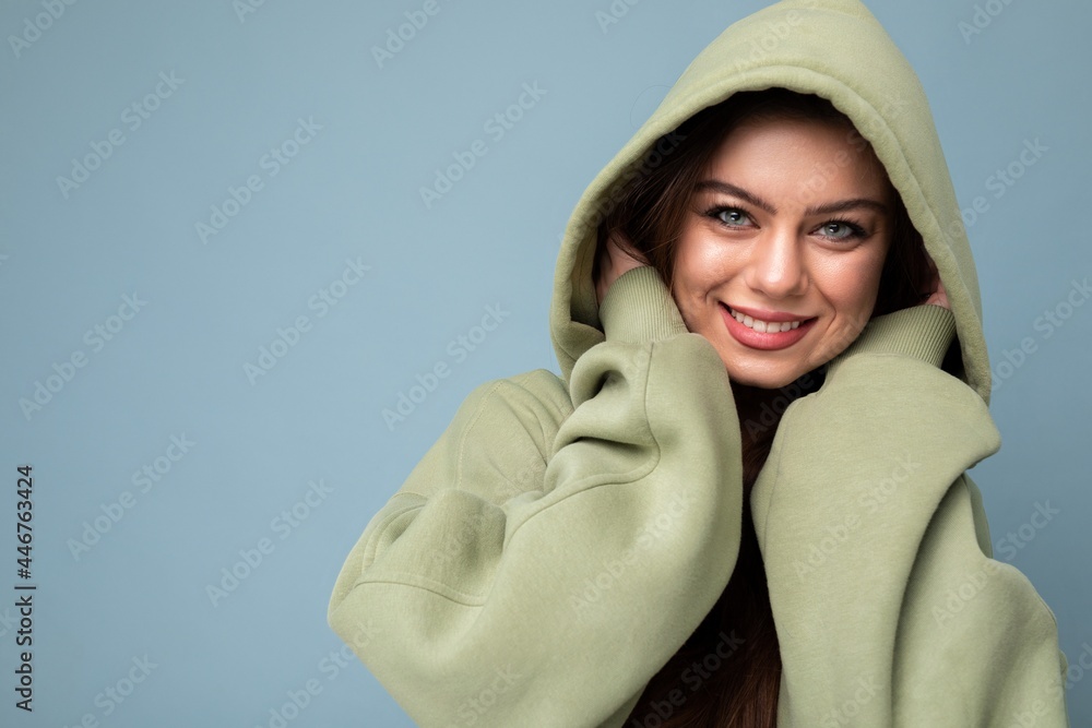 Portrait of attractive young caucasian smiling brunette woman model in trendy khaki hoodie and wearing hood isolated on blue background with copy space. Positive concept Portrait of attractive young caucasian smiling brunette woman model in trendy khaki hoodie and wearing hood isolated on blue background with copy space. Positive concept