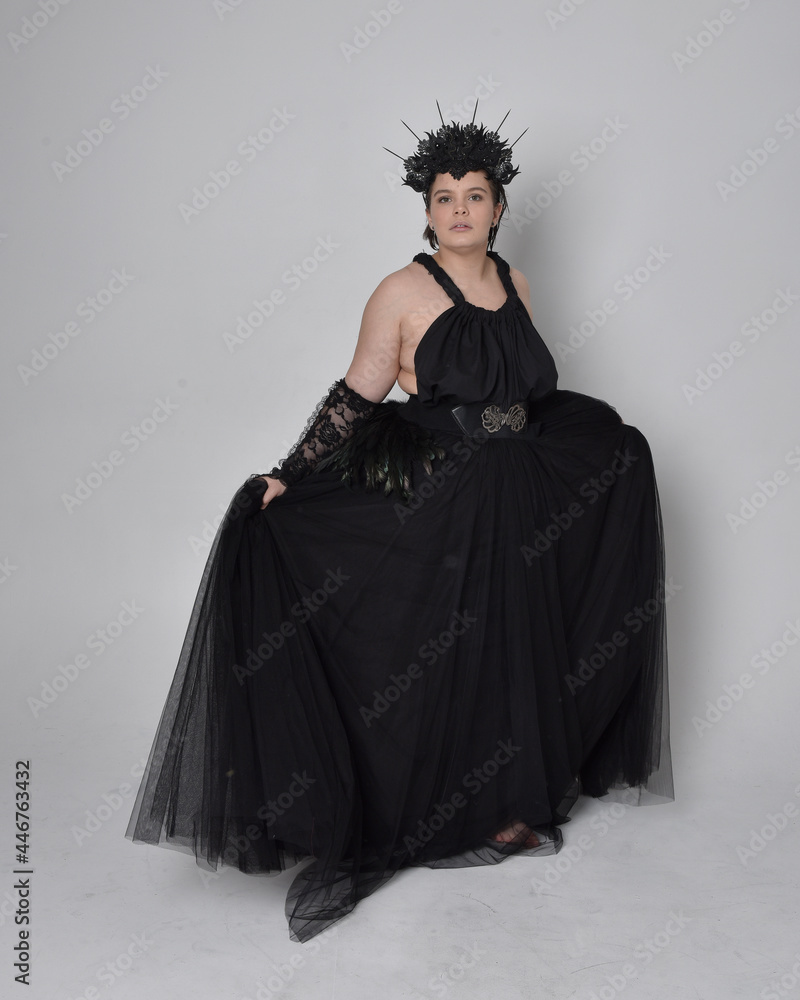 Naklejka premium Full length portrait of young plus sized woman with short hair, wearing long black tulle gothic gown with crown and gloves, standing pose on light studio background.