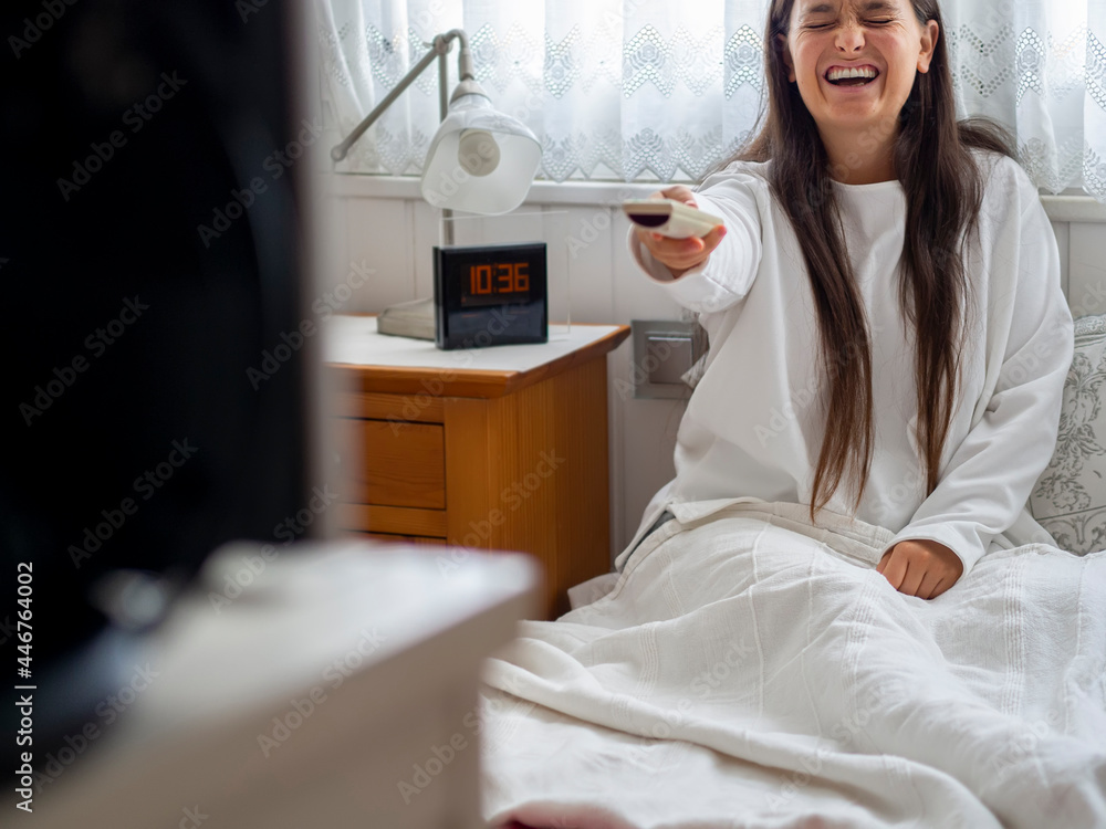 Woman laughing wearing pajamas sitting on bed watching TV. Relaxing woman in bedroom having a belly laugh watching a movie with remote control. Entertainment at home on the weekend, no plans concept