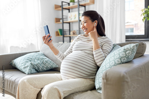 pregnancy, rest, people and expectation concept - happy smiling pregnant asian woman with smartphone and earphones sitting on sofa at home and listening to music