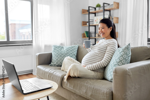 pregnancy, rest, people and expectation concept - happy smiling pregnant asian woman with laptop computer sitting on sofa at home and drinking tea
