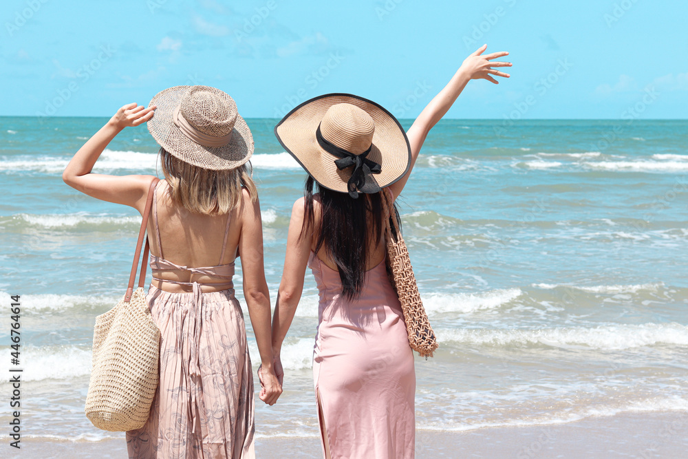 Poster Wandbilder Two beautiful Asian women with big hat from behind enjoy spending time on tropical sand beach blue sea together resting and