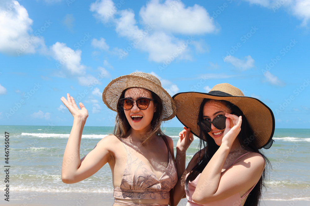 Two beautiful Asian women with big hat and sunglasses enjoy spending time with friend on tropical sand beach blue sea together, resting and relaxing on summer holiday vacation.