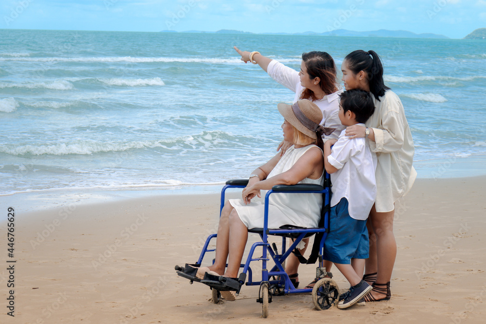 Happy disabled senior elderly woman in wheelchair spending time together with her family on tropical beach. Asian grandma, daughter and grandchildren resting and relaxing on summer holiday vacation