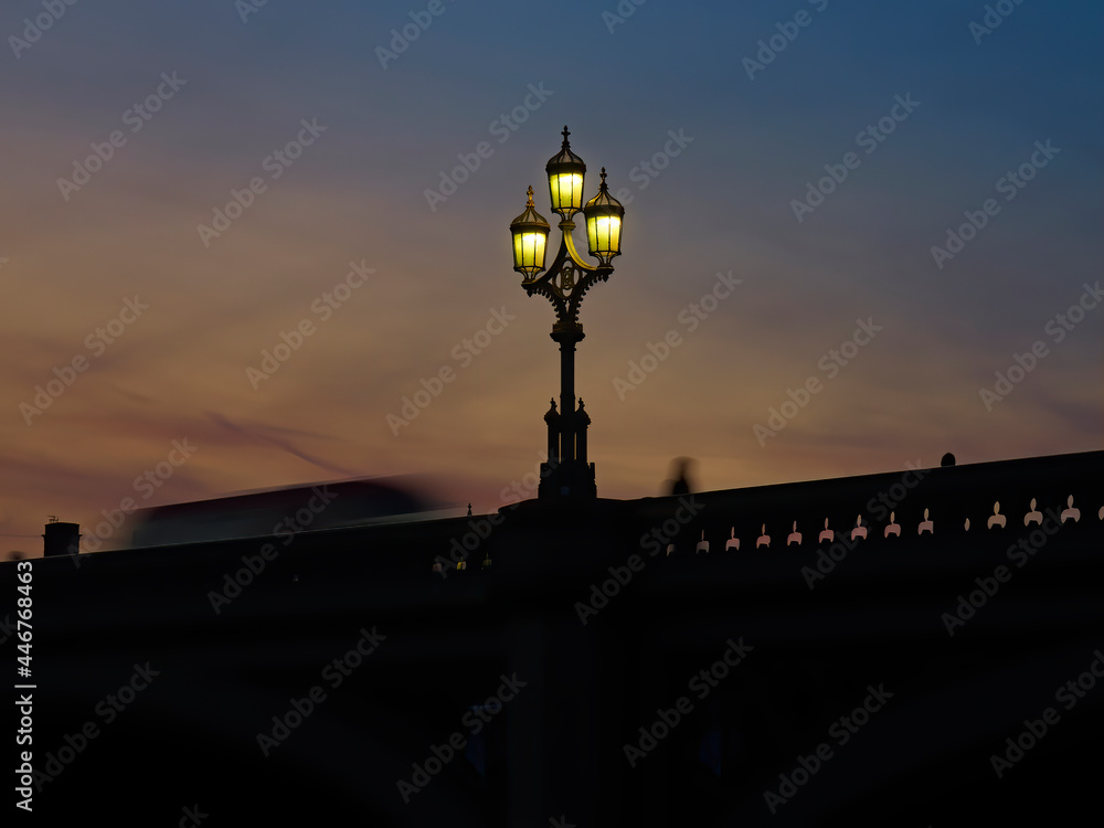 Low-key, soft view looking up to the parapet of Westminster Bridge where a gothic streetlight stands and a London Bus and person are silhouetted and motion-blurred ahead of the last embers of a sunset