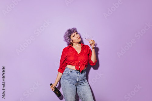 Cute wonderful girl in red stylish shirt holds glass and bottle of white wine and looks at camera on lilac isolated backdrop..