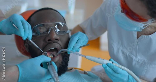 Close-up an African American male patient in eyeglasses is sitting in a dentist chair while the dentist is cleaning teeth by using a dental brush and mirror, female nurse is holding a saliva ejector.