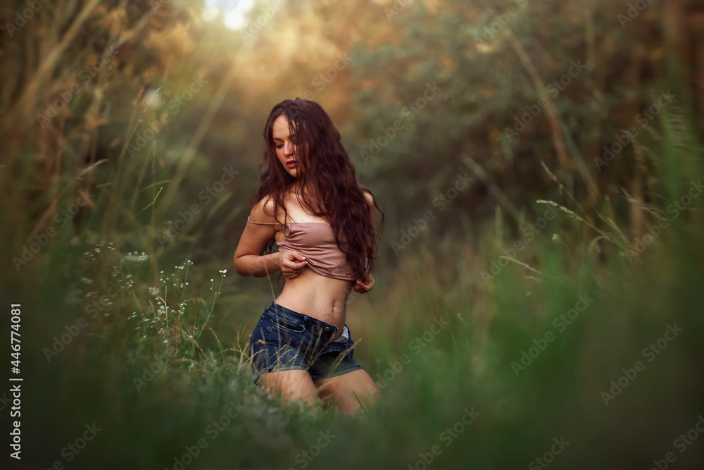 Sexy woman posing in a field in the grass rays of the sunset. Lifestyle on nature dressed in jeans shorts and beige top in golden hour. Romantic autumn mood, long tall grass in the meadow Sexy woman posing in a field in the grass rays of the sunset. Lifestyle on nature dressed in jeans shorts and beige top in golden hour. Romantic autumn mood, long tall grass in the meadow