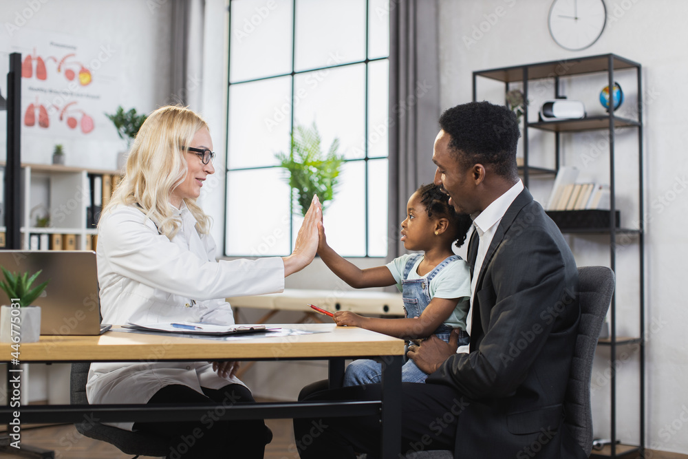 Pleasant caucasian pediatrician greeting little patient with high five at cabinet. African american father sitting at table with pretty daughter on knees. Regular checkup for children. Pleasant caucasian pediatrician greeting little patient with high five at cabinet. African american father sitting at table with pretty daughter on knees. Regular checkup for children.