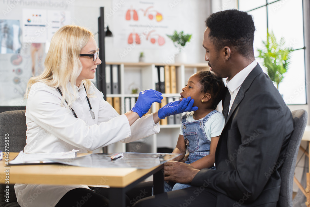 African american man holding his pretty daughter on knees while caucasian female pediatrician examining her throat. Concept of parenthood and health care. African american man holding his pretty daughter on knees while caucasian female pediatrician examining her throat. Concept of parenthood and health care.