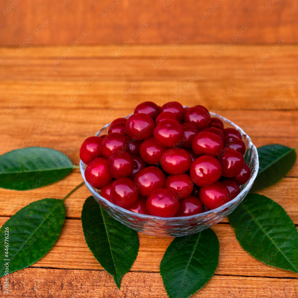 Ripe large juicy cherries in a transparent plate on a wooden table with green leaves. Ripe large juicy cherries in a transparent plate on a wooden table with green leaves.