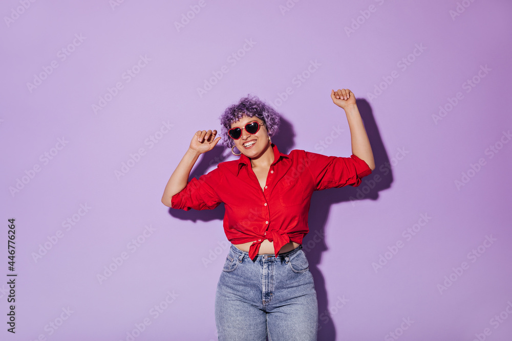 Curly-haired girl in sunglasses, red long-sleeved shirt and jeans smiles on purple background. Charming girl in stylish clothes posing for camera.. Curly-haired girl in sunglasses, red long-sleeved shirt and jeans smiles on purple background. Charming girl in stylish clothes posing for camera..