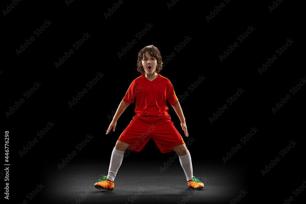 Happy, excited boy, football soccer player in red sports uniform with football ball isolated on dark studio background. Concept of sport, game, hobby