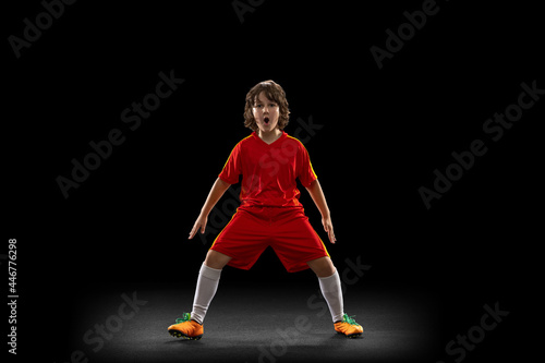Happy, excited boy, football soccer player in red sports uniform with football ball isolated on dark studio background. Concept of sport, game, hobby