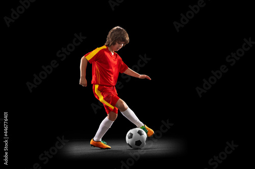 In motion. One little male football soccer player, boy training with football ball isolated on dark studio background. Concept of sport, game, hobby © master1305