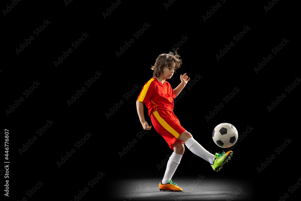 One little male football soccer player, boy training with football ball isolated on dark studio background. Concept of sport, game, hobby