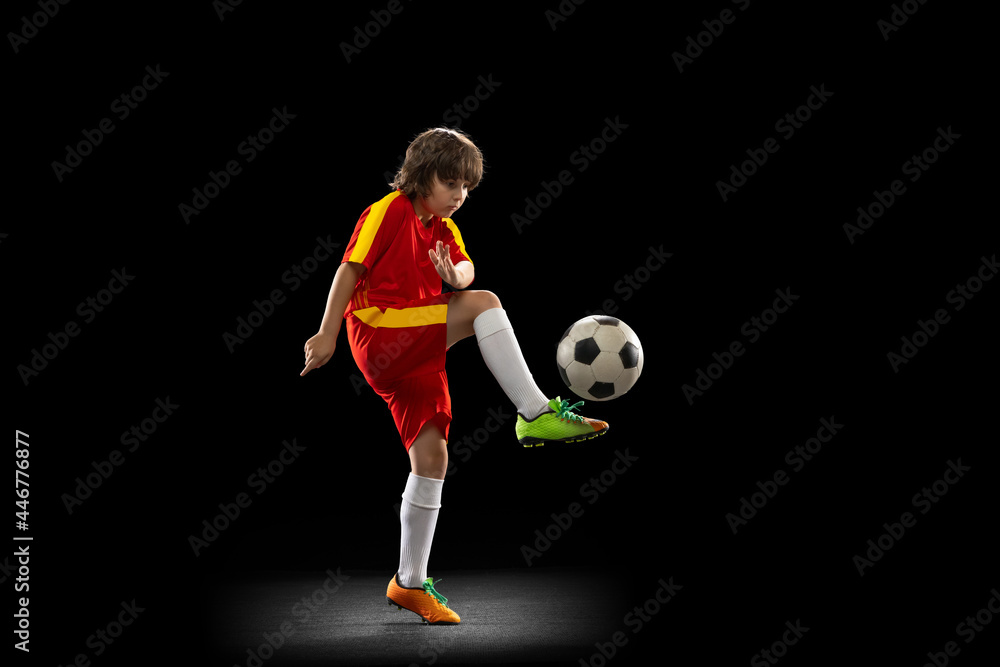 In motion. One little male football soccer player, boy training with football ball isolated on dark studio background. Concept of sport, game, hobby