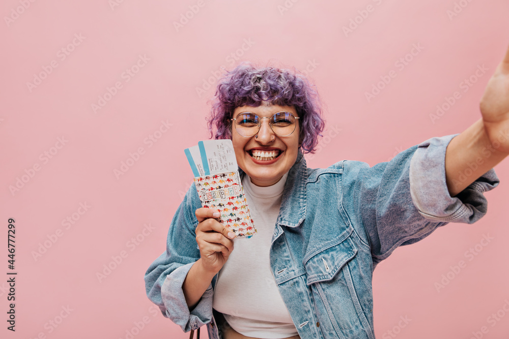 Happy girl with purple short curly hair in round glasses, blue jacket and white top sincerely smiles on pink background.. Happy girl with purple short curly hair in round glasses, blue jacket and white top sincerely smiles on pink background..