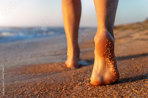 Close up detail bottom behind pov view of pretty female person barefoot legs heel walking by scenic sandy ocean or sea beach at warm golden sunrise morning. Wild nature travel and vacation concept