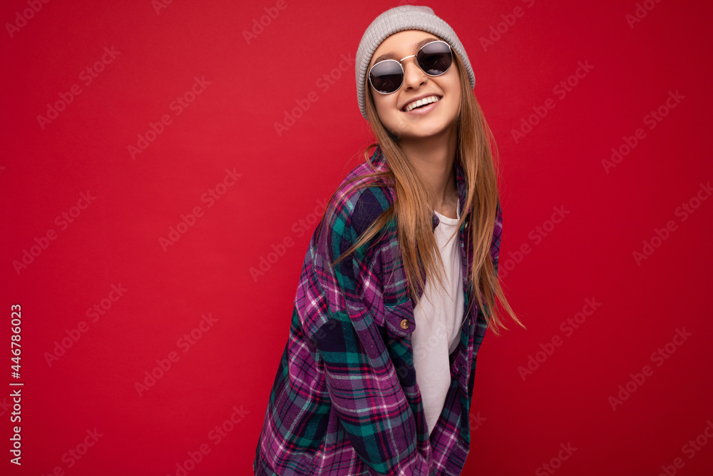 Fascinating happy young dark blonde woman isolated over red background wearing purple casual shirt grey hat and stylish sunglasses looking at camera and smiling