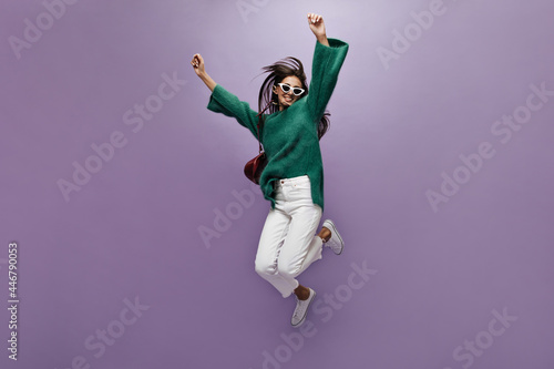 Fototapeta Naklejka Na Ścianę i Meble -  Tanned joyful woman in stylish white pants, green sweater and sunglasses jumps on isolated. Brunette long-haired girl moves on purple background.