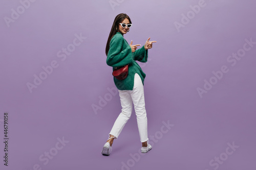 Stylish woman in sunglasses, green sweaterwhite pants and sneakers with claret handbag dances on purple background. Happy girl moves, also points at some place