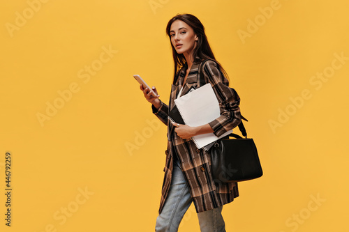 Young brunette woman in oversized jacket and jeans holds phone and white notebooks. Attractive girl with black bag poses on orange background.