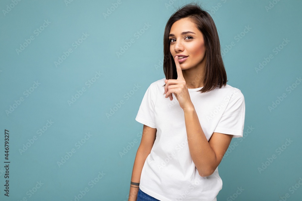 Portrait of young attractive brunette woman with sincere emotions wearing casual white t-shirt for mockup isolated on blue background with copy space and showing silence gesture with finger near lips Portrait of young attractive brunette woman with sincere emotions wearing casual white t-shirt for mockup isolated on blue background with copy space and showing silence gesture with finger near lips