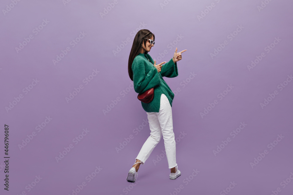 Pretty woman in stylish glasses, green woolen sweater and white pants points at place for text on purple background. Cheerful girl stands with her back but turned to the right