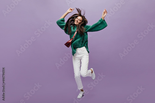 Thin tanned woman in white pants and green woolen sweater dances on purple background. Charming girl with cross-body bag jumps and moves on isolated. © Look!