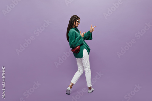 Pretty woman in stylish glasses, green woolen sweater and white pants points at place for text on purple background. Cheerful girl stands with her back but turned to the right