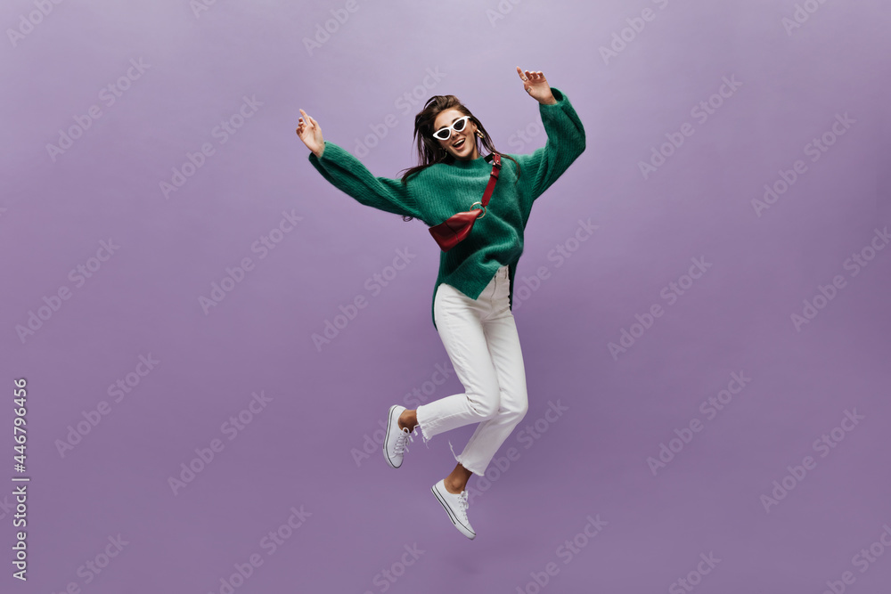 Cheerful woman in white pants and stylish oversized green sweater moves on purple background. Happy brunette girl in sunglasses jumps on isolated. Cheerful woman in white pants and stylish oversized green sweater moves on purple background. Happy brunette girl in sunglasses jumps on isolated.