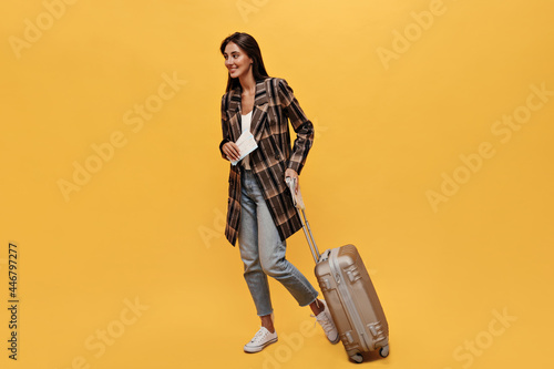 Brunette long-haired woman in denim pants and brown coat poses with tickets and suit-bag. Portrait of happy travel girl moves and smiles on isolated yellow background.