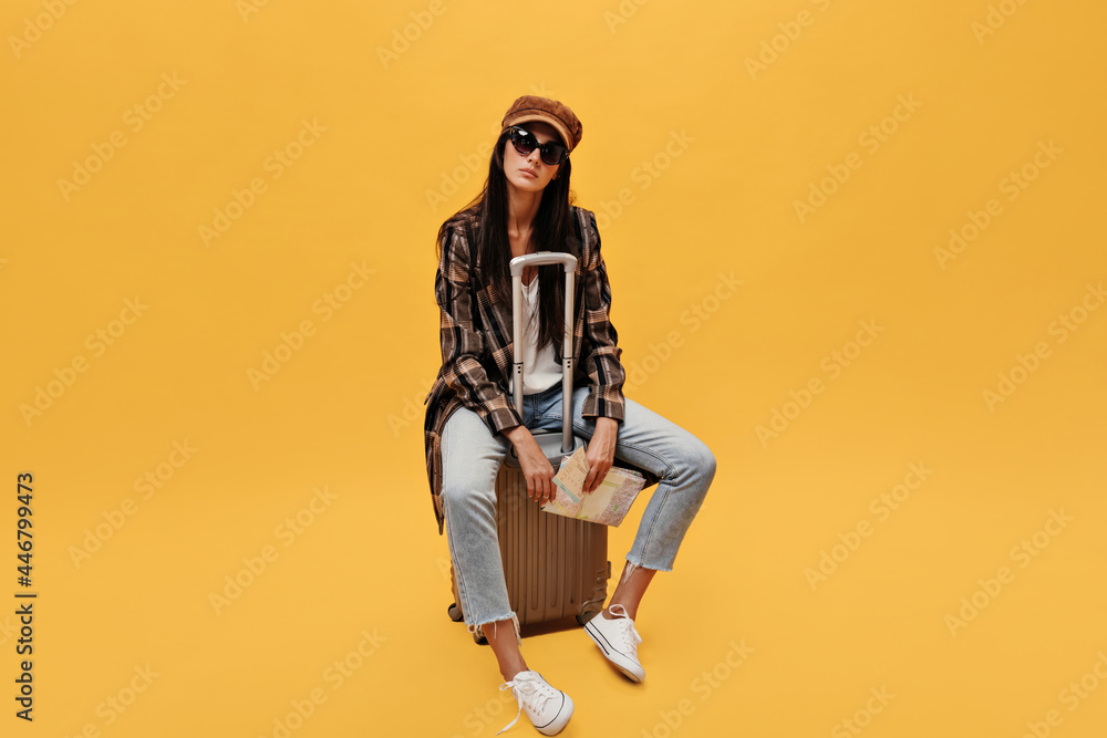 Serious woman in brown beret and sunglasses sits on suit bag. Brunette girl in coat and jeans holds tickets on isolated yellow background. Serious woman in brown beret and sunglasses sits on suit bag. Brunette girl in coat and jeans holds tickets on isolated yellow background.